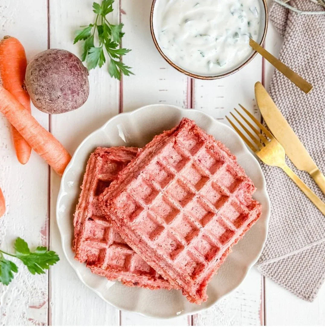 Pink waffles on a white plate with carrots, herbs, and a bowl of sauce on a wooden surface.