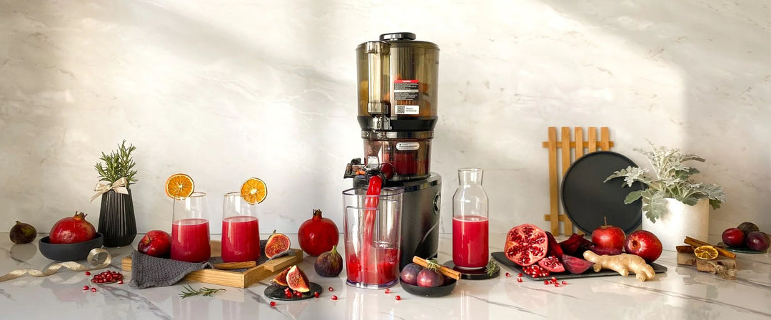 Kuvings AUTO10S black slow juicer making fresh pomegranate juice with fruits and juice glasses on a kitchen countertop.