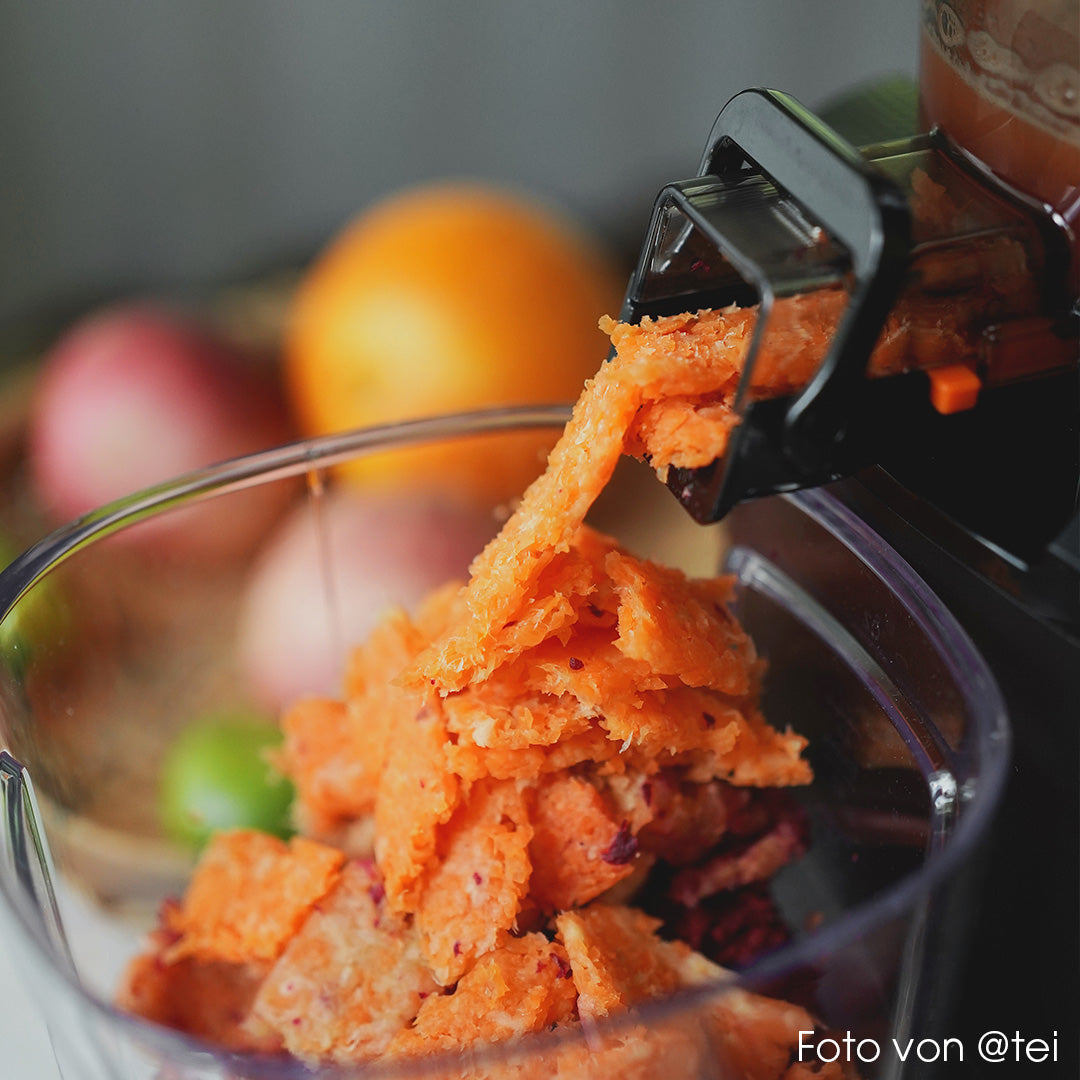 Close-up lifestyle shot of a Kuvings AUTO10S slow juicer ejecting dry orange carrot pulp into a clear container, with fresh fruit blurred in the background.