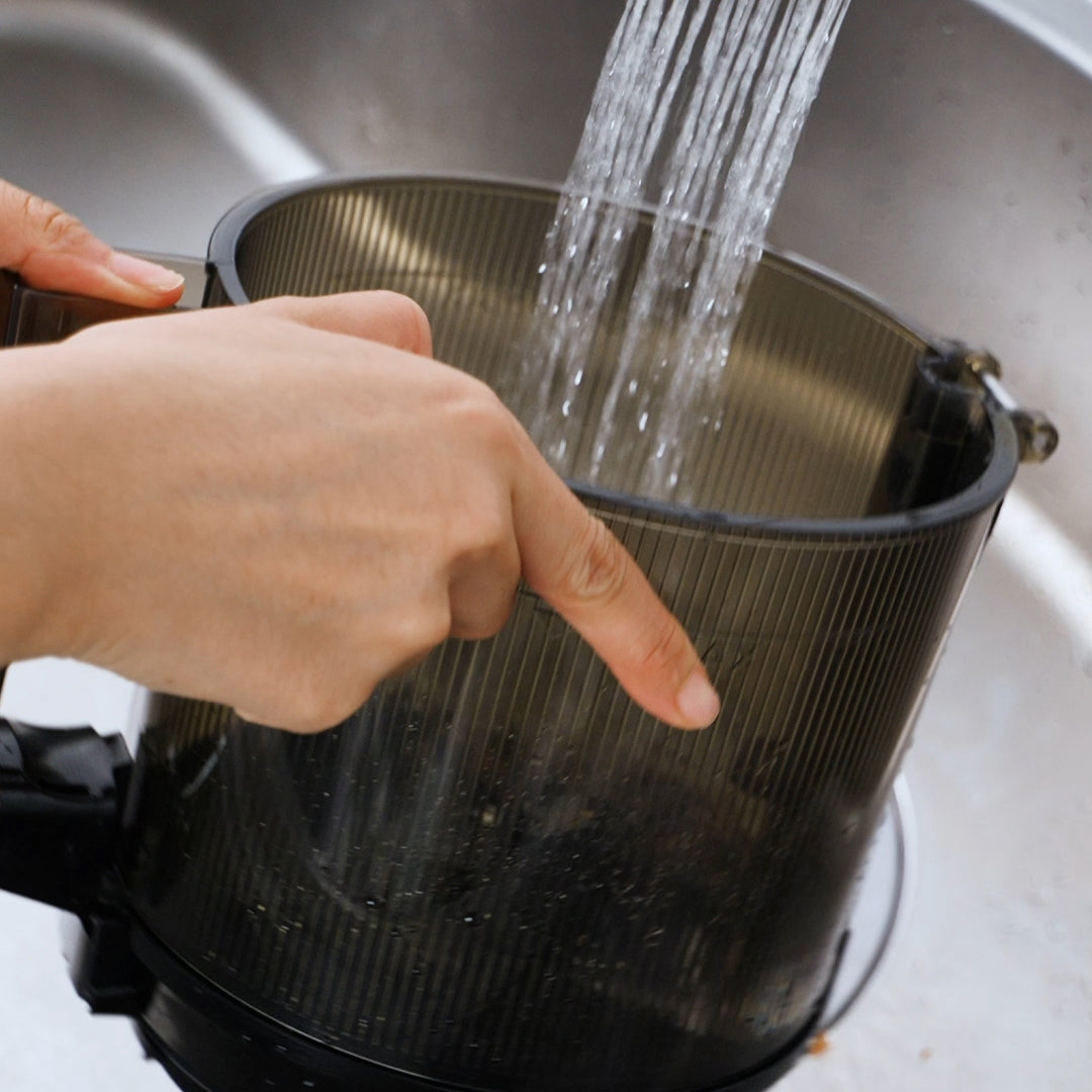 Close-up lifestyle shot of the transparent grey Kuvings AUTO10S mega-hopper container being rinsed under a stream of running tap water in a kitchen sink.