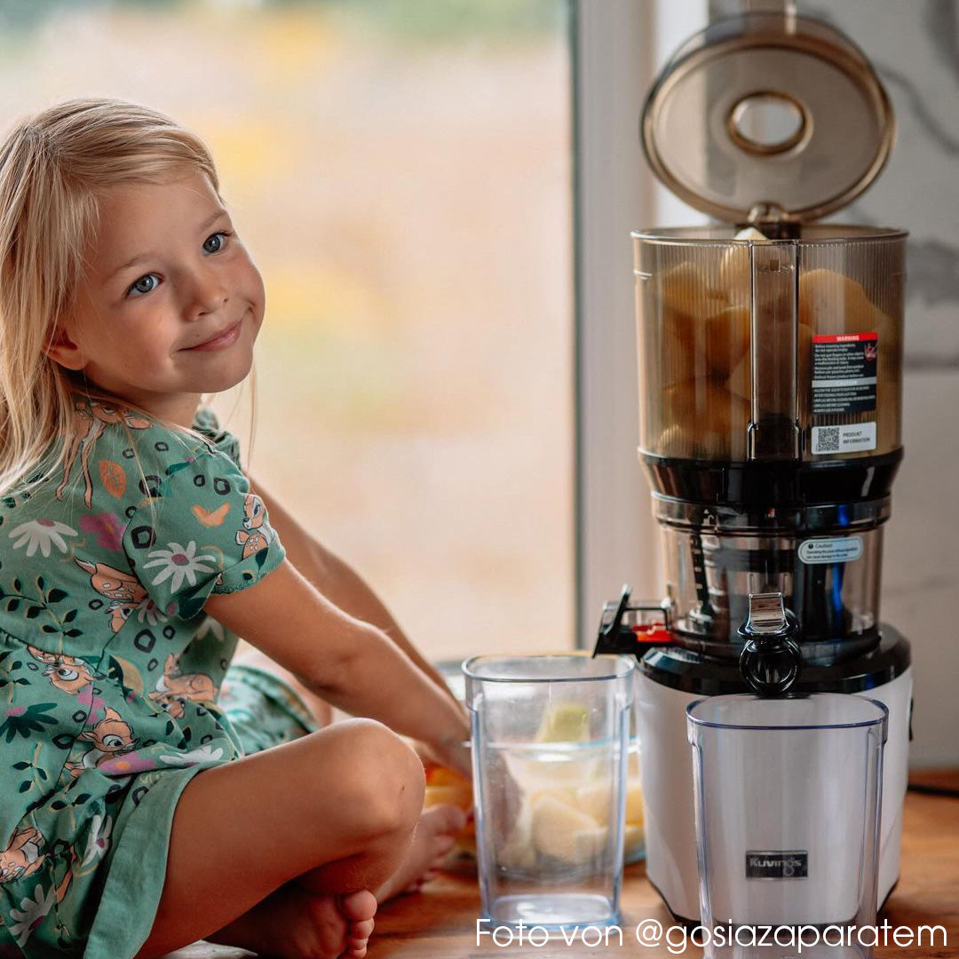 Lifestyle shot of a little girl sitting next to a quiet white Kuvings AUTO10S slow juicer filled with chopped ingredients, with German photo credit text at the bottom.
