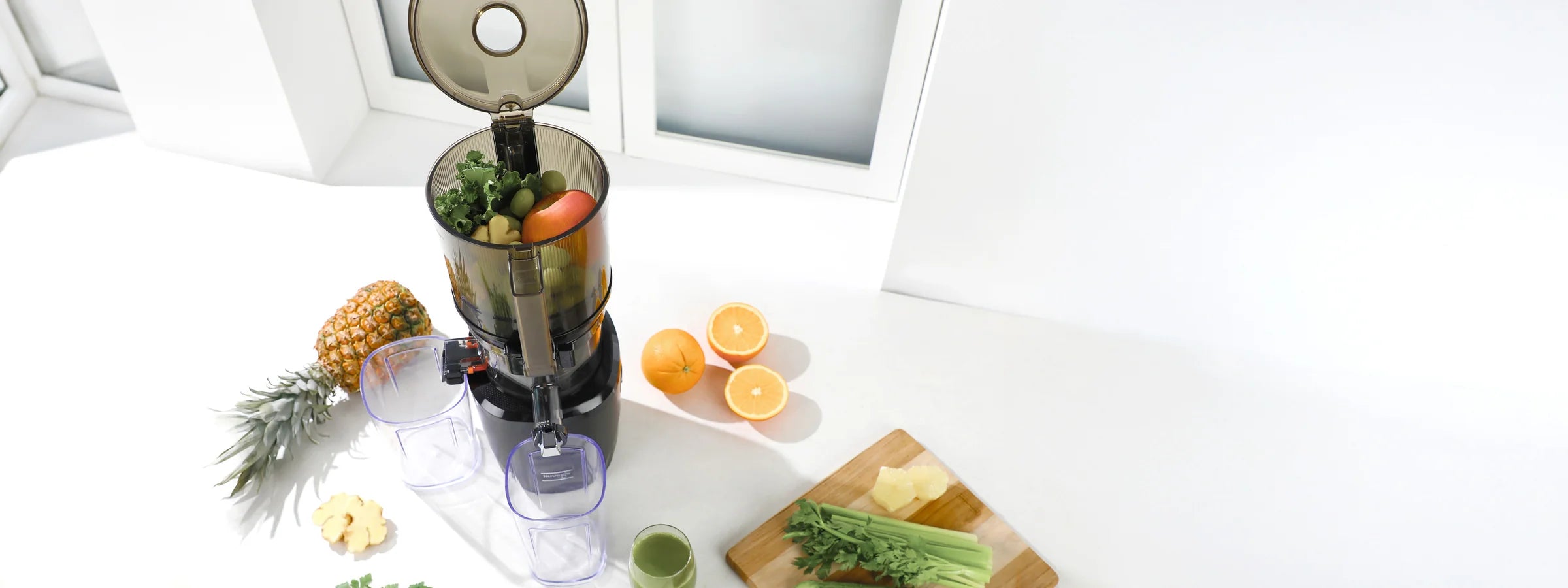 High-angle panoramic banner of an open black Kuvings AUTO10S slow juicer filled with fruits and greens on a white countertop, surrounded by a whole pineapple, halved oranges, ginger, and fresh celery.