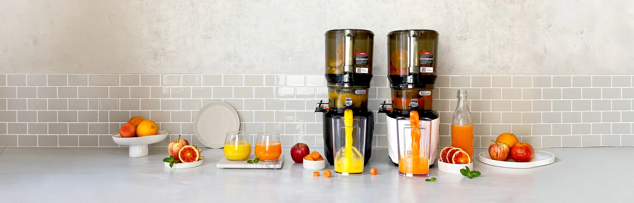 Panoramic banner of a black and a white Kuvings AUTO10S slow juicer side-by-side extracting fresh orange and carrot juice into pitchers on a kitchen counter.
