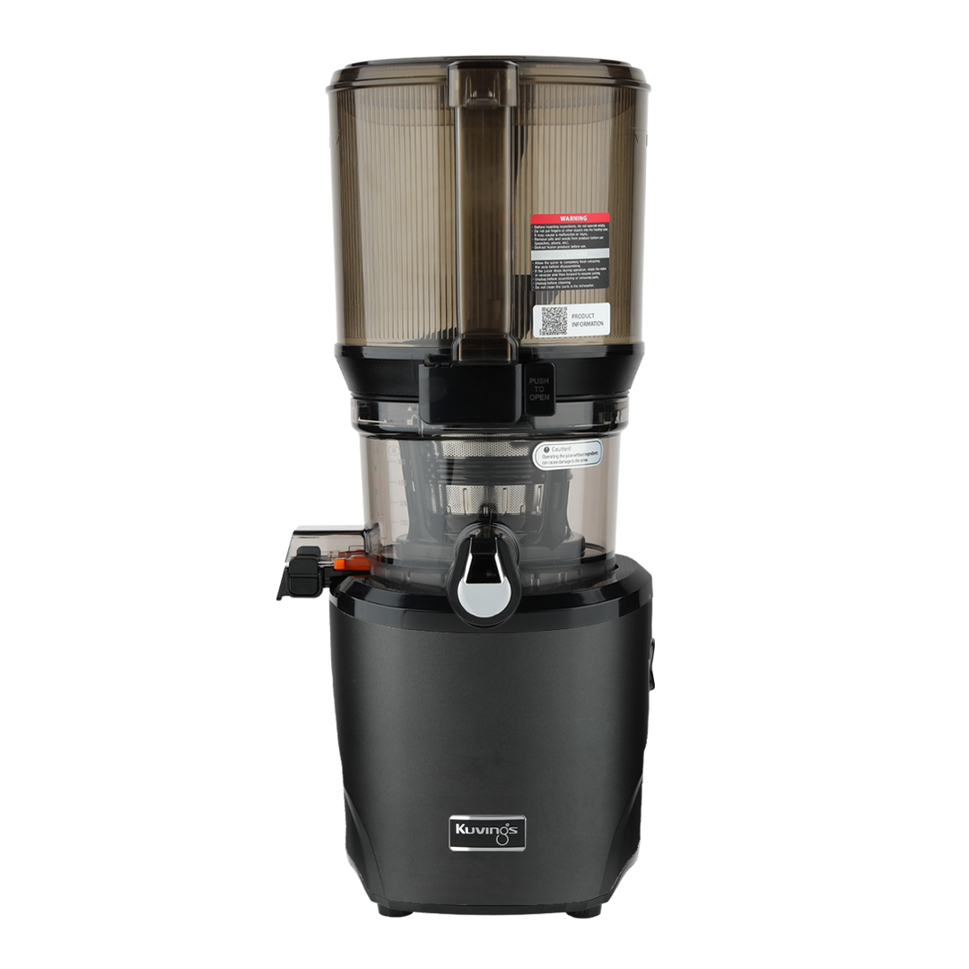 Straight-on studio product shot of a matte black Kuvings AUTO10S whole slow juicer with its large transparent grey mega-hopper container attached, showing the safety warning labels and QR code sticker, isolated against a transparent grid background.