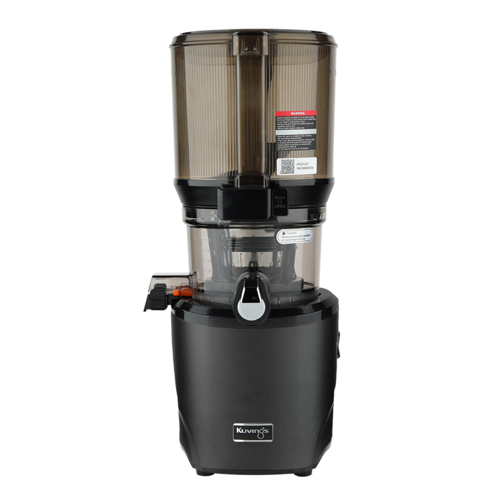 Straight-on studio product shot of a matte black Kuvings AUTO10S whole slow juicer with its large transparent grey mega-hopper container attached, showing the safety warning labels and QR code sticker, isolated against a transparent grid background.
