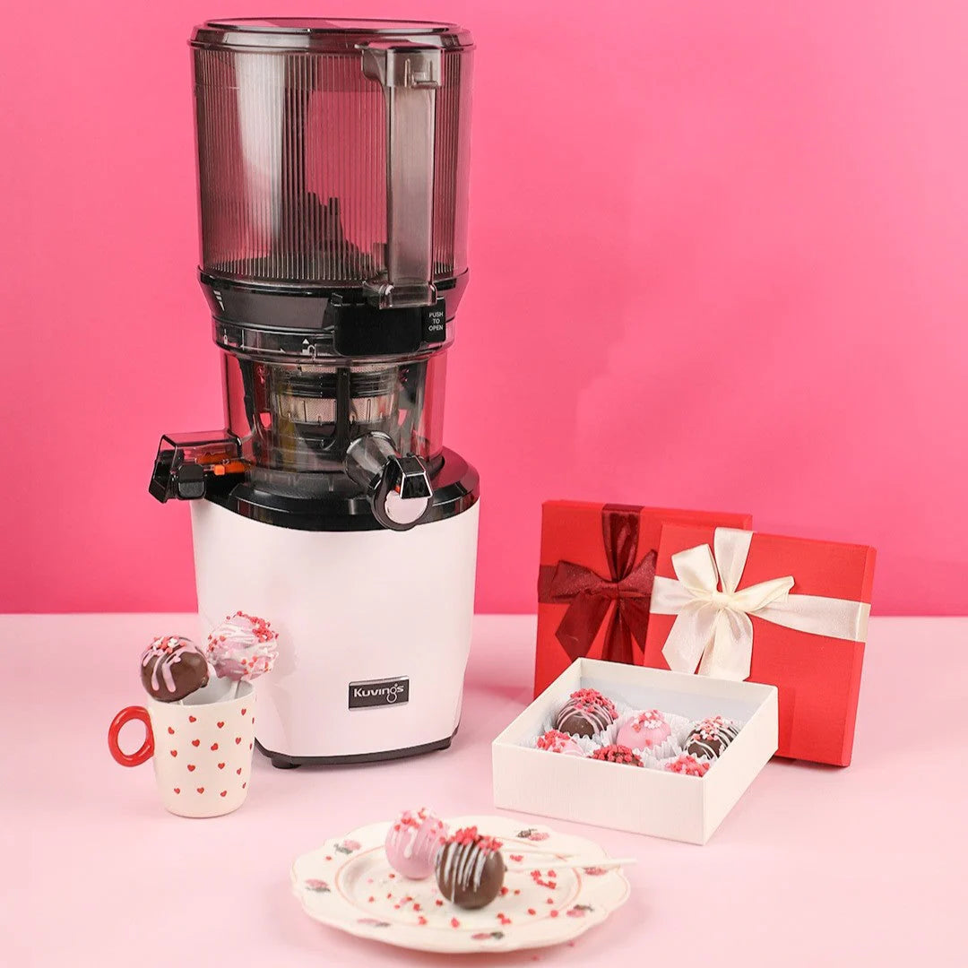 Studio product shot of a white Kuvings AUTO10S slow juicer arranged for Valentine's Day with wrapped red gift boxes, a white box of decorated cake pops, a heart-patterned mug, and a matching dessert plate against a vibrant pink background.