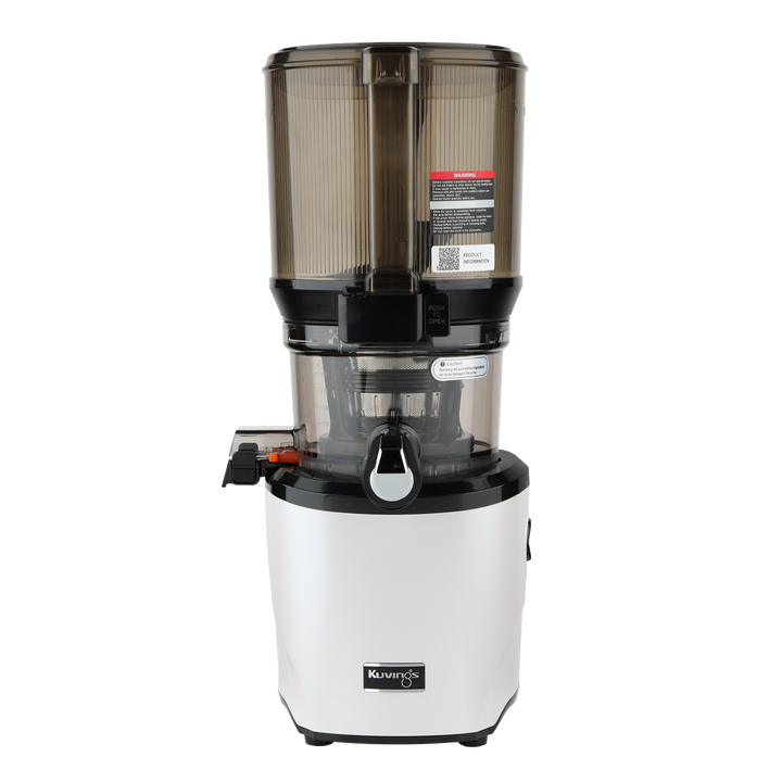 Straight-on studio product shot of a white Kuvings AUTO10S whole slow juicer with its large transparent grey mega-hopper container attached, showing the closed smart cap spout and safety labels, isolated against a transparent grid background.