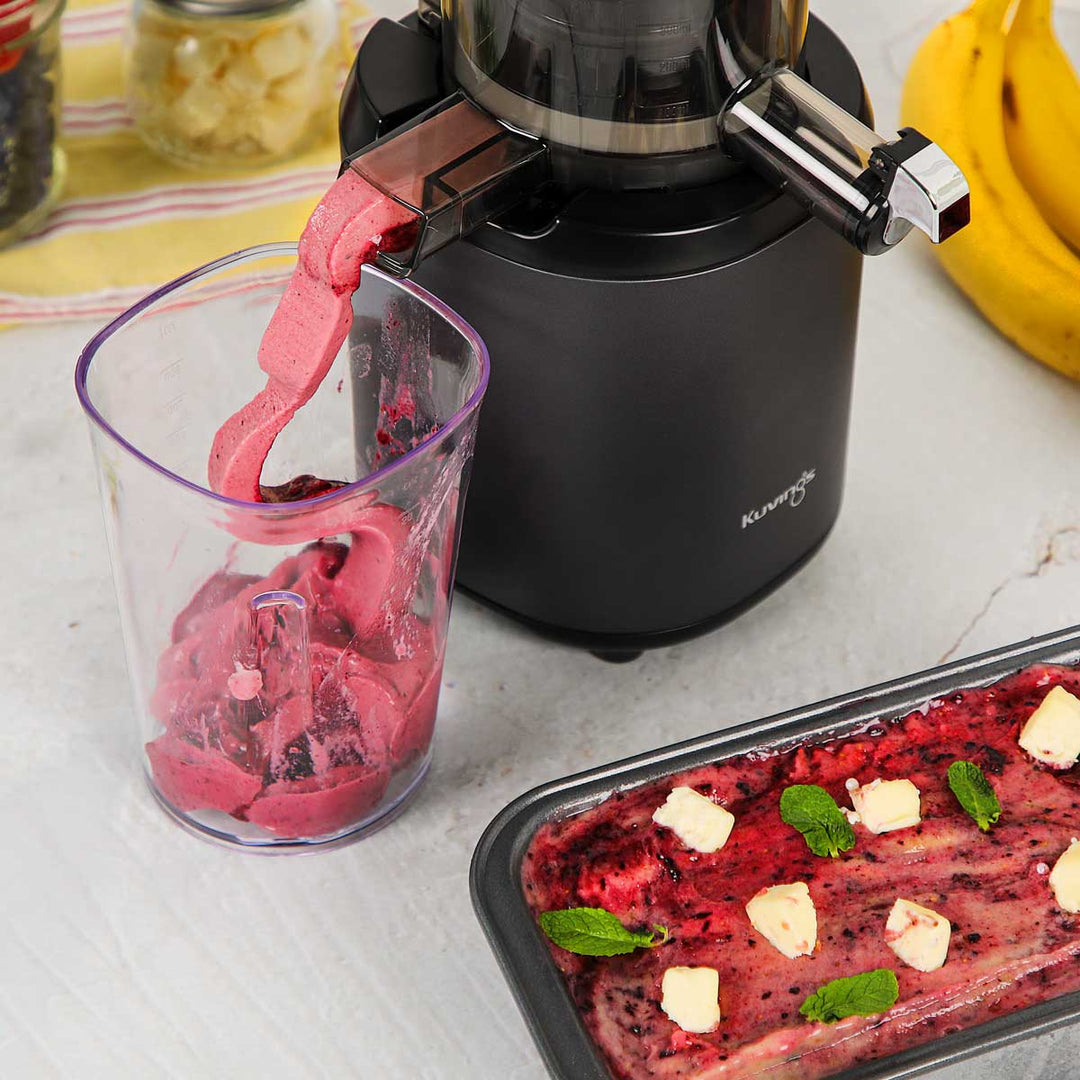 Close-up shot of a matte black Kuvings AUTO6 slow juicer dispensing fresh berry sorbet into a clear container, alongside a tray of decorated fruit ice cream.