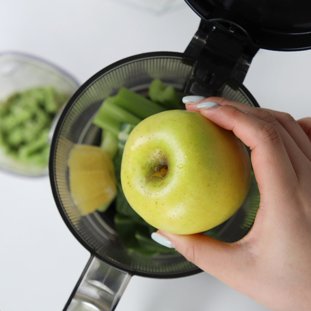Close-up shot of a hand placing a whole green apple into the open hopper of a Kuvings AUTO6 slow juicer filled with celery stalks and leafy greens.