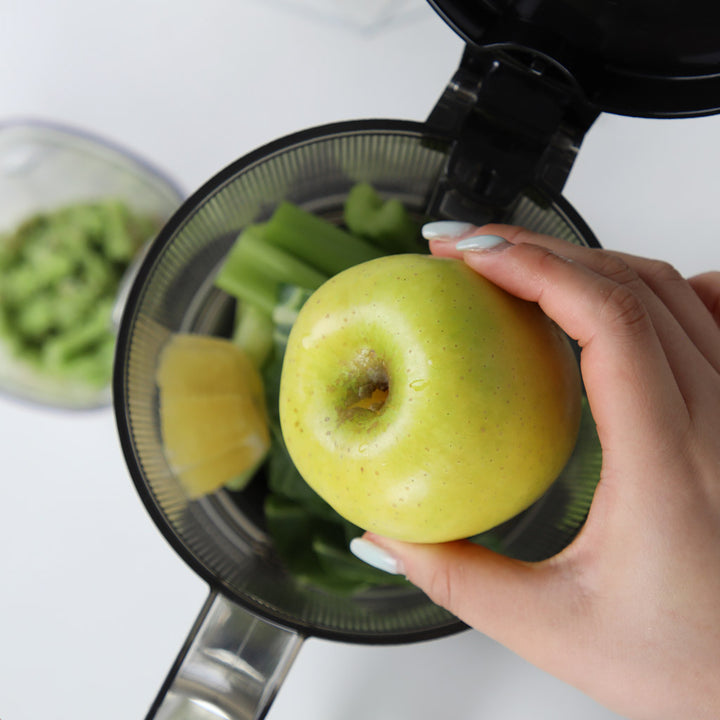 Close-up shot of a hand placing a whole green apple into the open hopper of a Kuvings AUTO6 slow juicer filled with celery stalks and leafy greens.