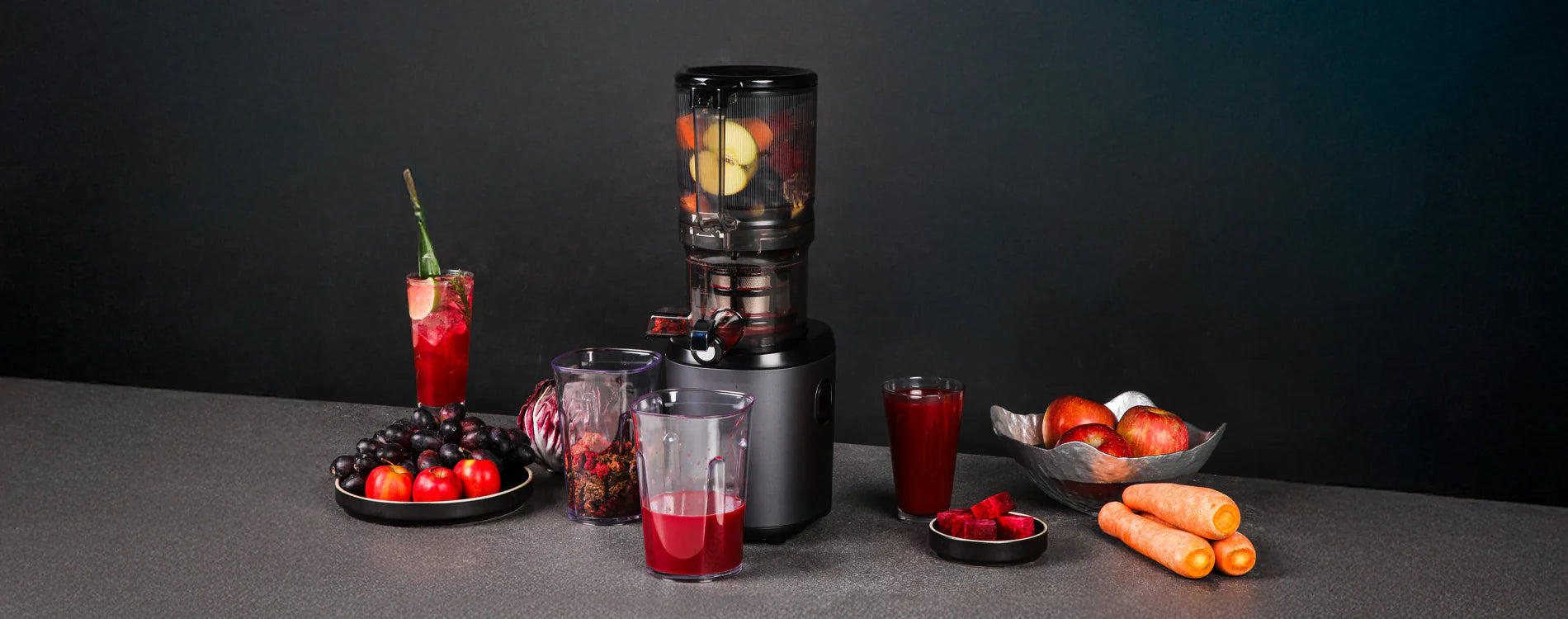 Panoramic lifestyle banner shot of a graphite grey Kuvings AUTO6 juicer with whole apples and carrots inside its mega-hopper, surrounded by pitchers of red juice, fresh produce, and a cocktail glass on a dark counter.