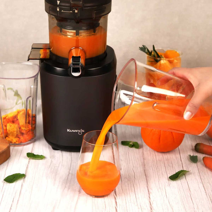 Close-up lifestyle shot of a hand pouring freshly pressed orange carrot juice from a clear pitcher into a glass next to a matte black Kuvings AUTO6 slow juicer.