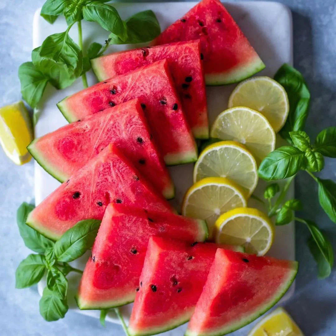 Slices of watermelon with lemon wedges and basil leaves on a white plate.