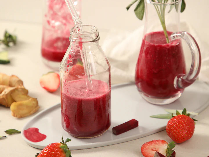 Red smoothie in a glass bottle and pitcher with strawberries and other fruits on a white surface.