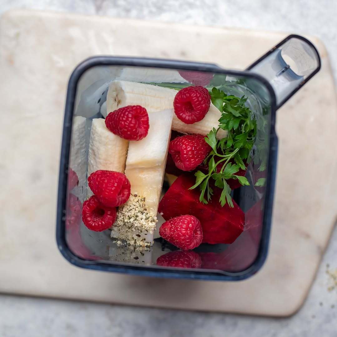 Top-down, bird's-eye view looking inside a Kuvings blender jar filled with fresh smoothie ingredients, including sliced bananas, red raspberries, beetroot pieces, green parsley, and hemp seeds, resting on a marble cutting board.