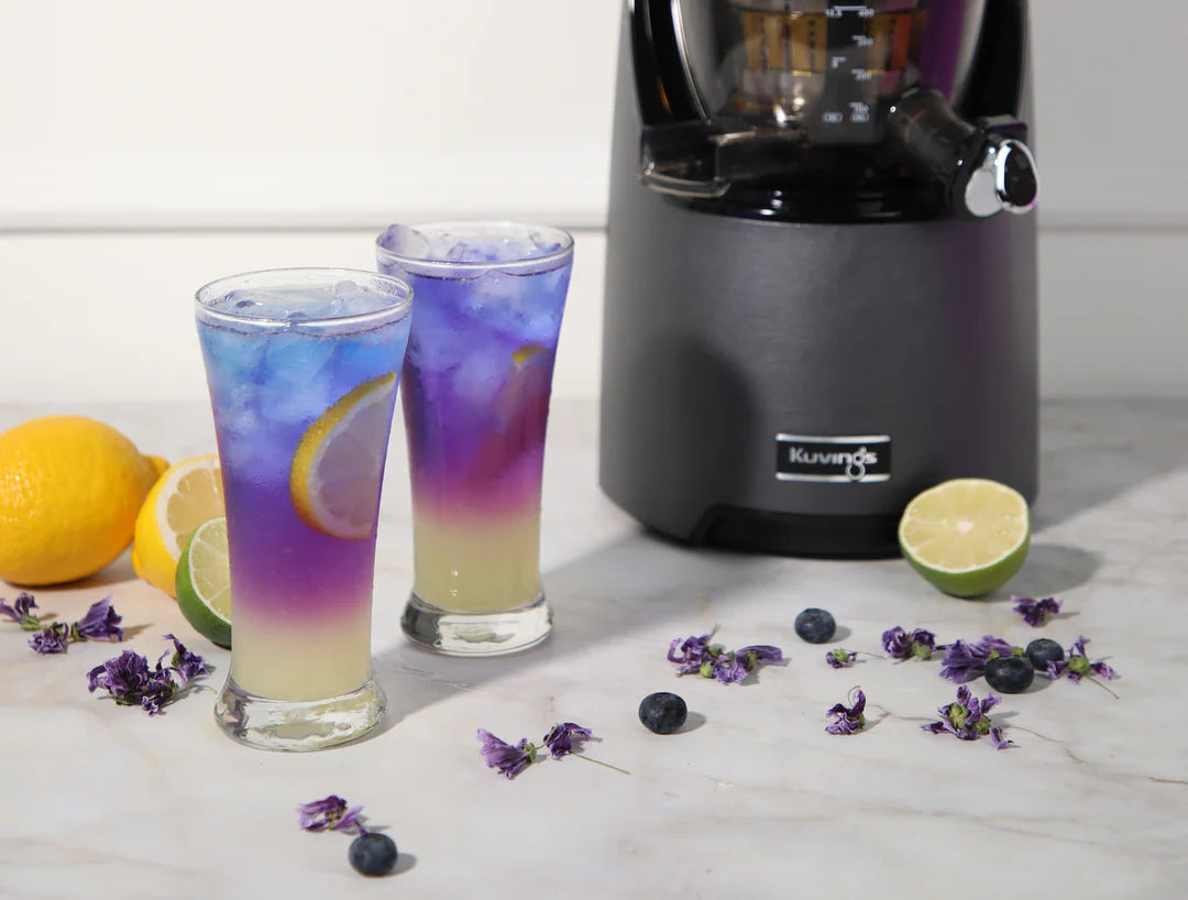 Two colorful layered drinks in glasses with a juicer and fruits on a marble surface with black kuvings machine in the background.