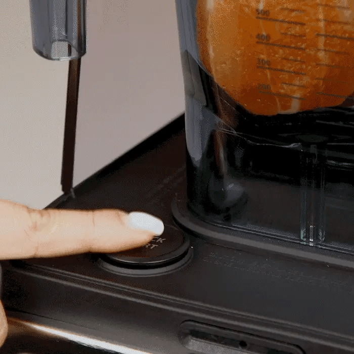 Close-up view of a finger with white nail polish pressing the quick start button on the base of a Kuvings CB1000 blender, next to a blending jar filled with orange fruit.