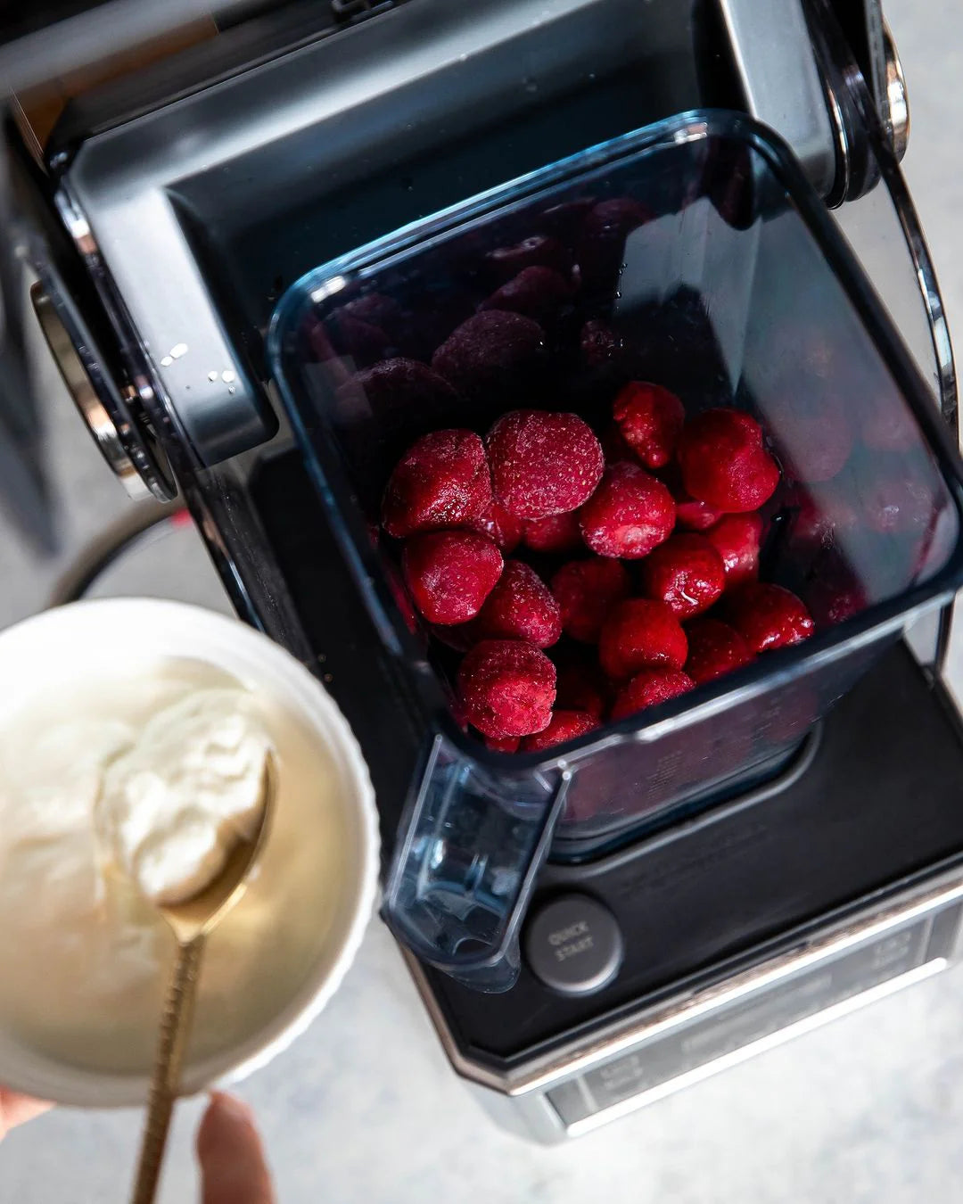 High-angle view of a Kuvings CB1000 blending jar filled with frozen red strawberries, positioned on the motor base next to a white bowl of thick yogurt with a gold spoon.