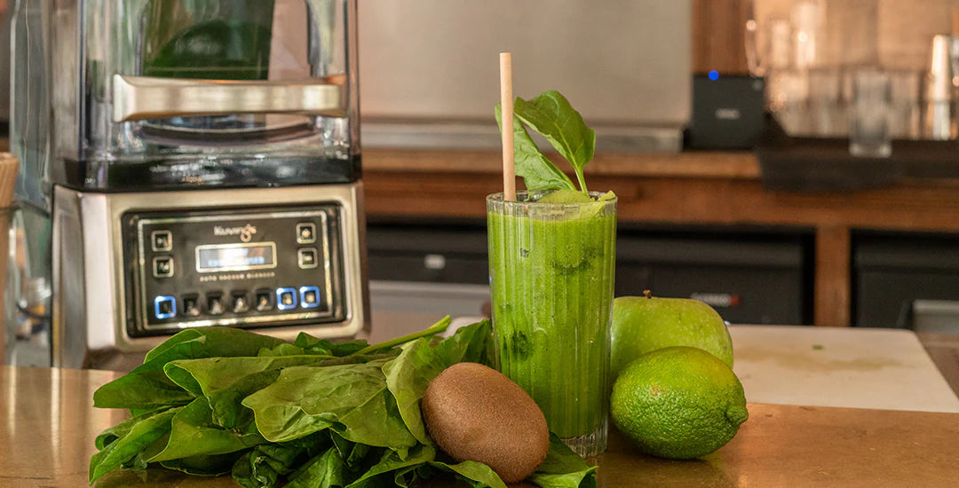 Breite Aufnahme auf Augenhöhe eines frischen grünen Smoothies in einem hohen Glas, umgeben von Spinatblättern, Kiwi, Limette und einem grünen Apfel auf einer Arbeitsplatte, mit einem Kuvings CB1000 gewerblichen Vakuum-Standmixer im Hintergrund.