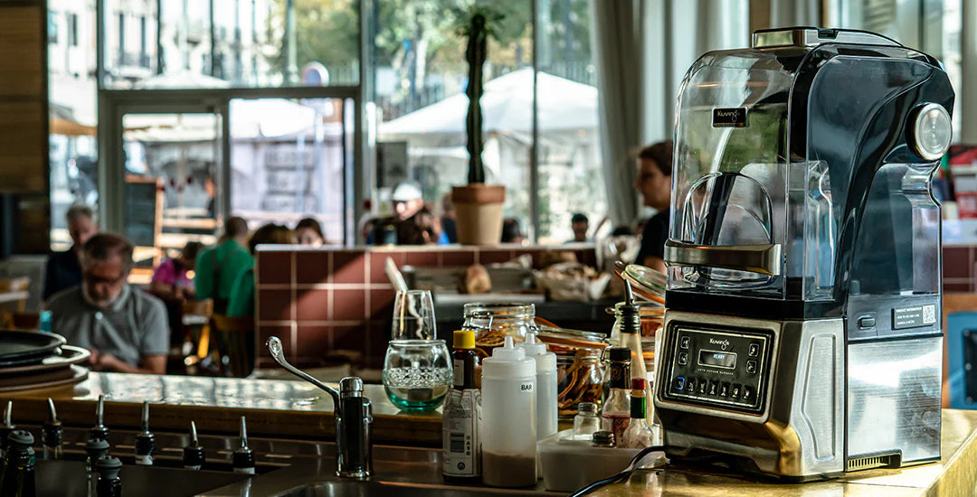 Angled eye-level view of a Kuvings CB1000 professional vacuum blender with its sound enclosure closed, standing on a cafe bar counter with a blurred background of a busy restaurant interior.