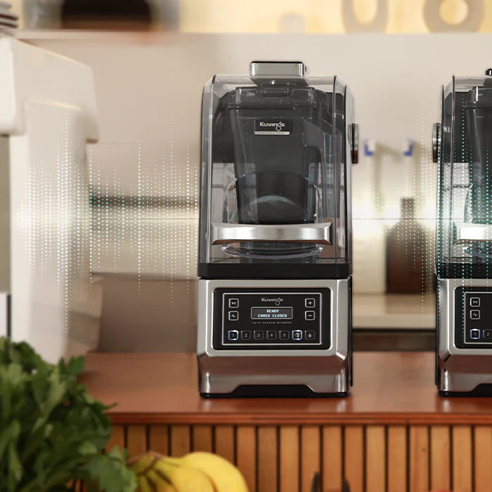 Straight-on, eye-level view of a Kuvings CB1000 professional auto vacuum blender on a wooden counter, featuring a horizontal soundwave graphic overlay to emphasize quiet operation, against a warm kitchen background.