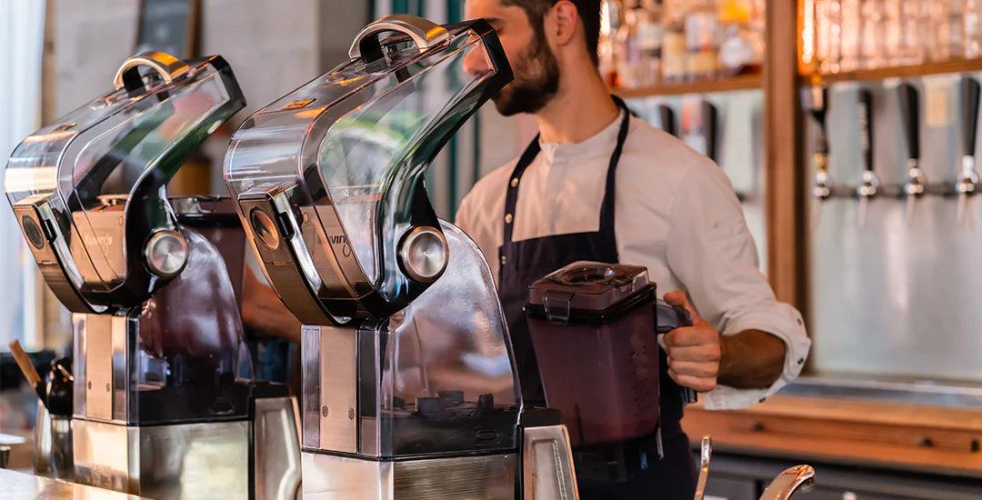Eye-level medium shot of two Kuvings CB1000 commercial vacuum blenders with their sound shields open on a bar counter, with a male barista in an apron holding a blending jar in the background.