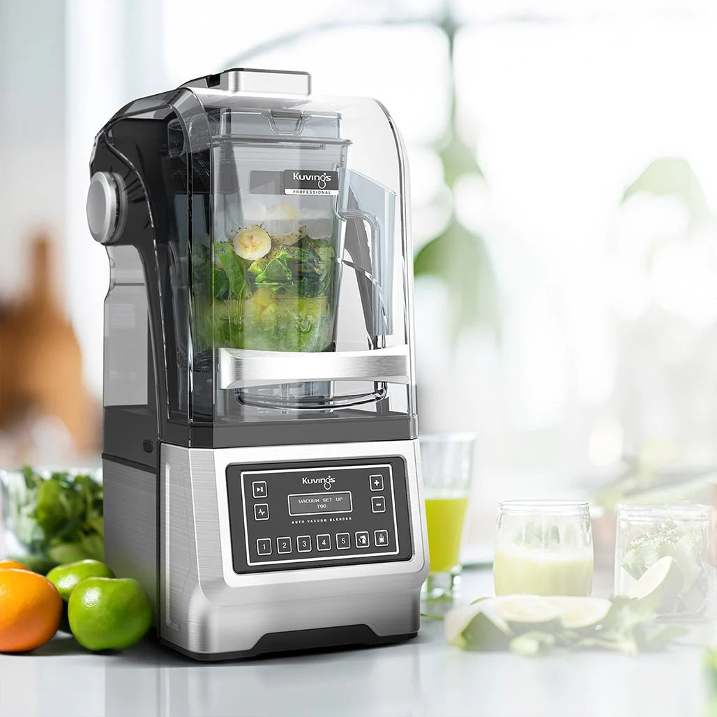 Angled eye-level shot of the Kuvings CB1000 Professional Auto Vacuum Blender with its clear sound enclosure closed, blending a green smoothie containing spinach and banana slices on a bright kitchen counter surrounded by fresh fruits.