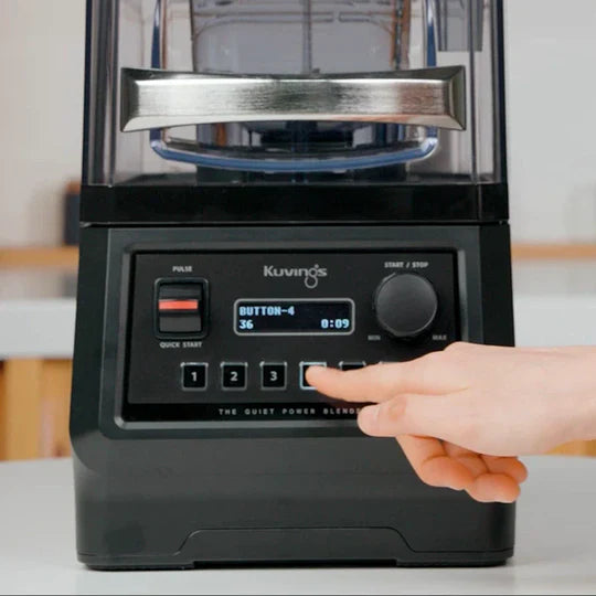 Straight-on, close-up front view of the Kuvings CB850 Quiet Power Blender control panel, showing a person's hand pressing a pre-programmed menu button with the digital screen displaying 'BUTTON-4' in a kitchen setting.