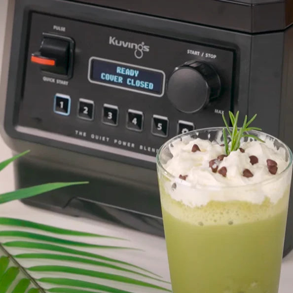 Angled close-up view of a fresh green tea smoothie topped with whipped cream and chocolate chips, placed in front of a Kuvings CB850 blender displaying 'READY COVER CLOSED' on its digital screen.