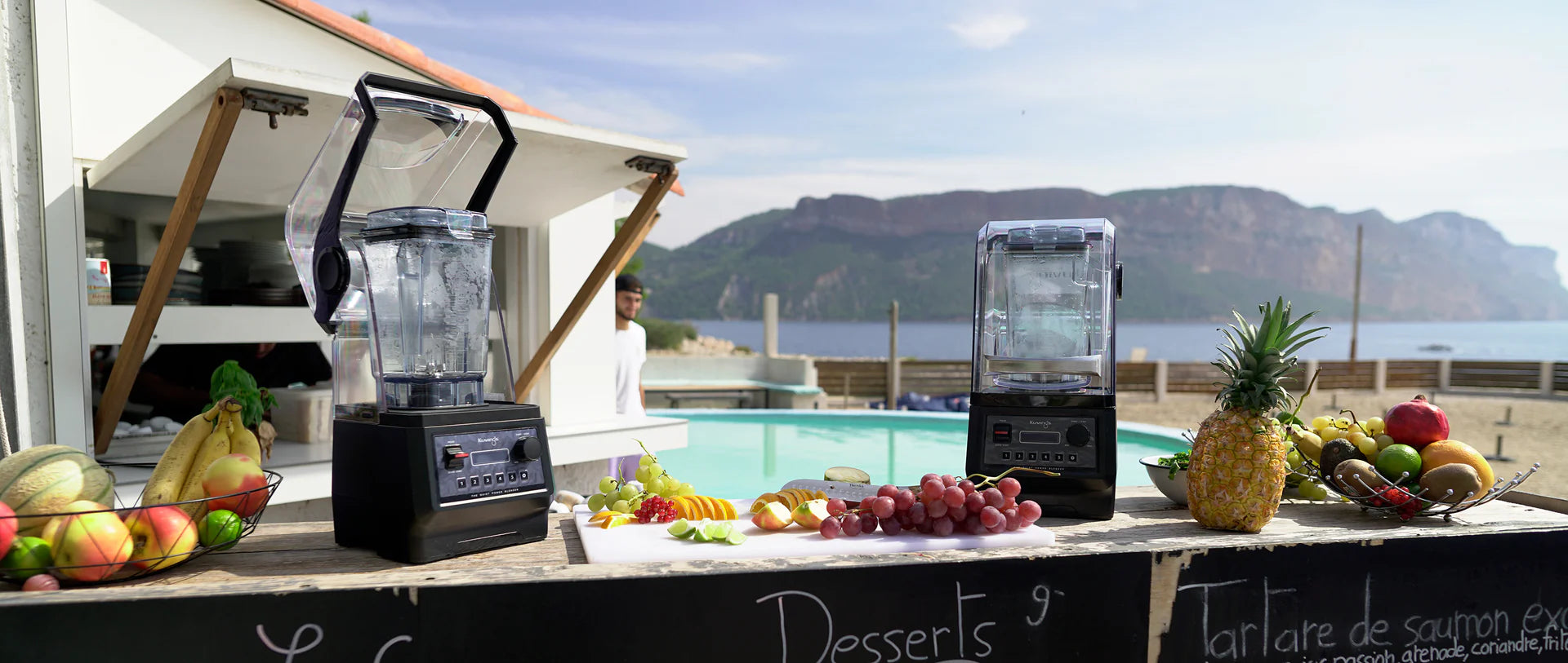 Eye-level wide lifestyle view of two Kuvings CB850 commercial blenders on an outdoor wooden bar counter surrounded by fresh fruits, with a swimming pool, ocean, and mountains in the background.