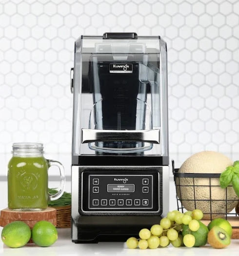 Straight-on, eye-level front view of a Kuvings CB980 professional auto blender with its clear sound enclosure closed, standing on a kitchen counter next to a mason jar of green juice and fresh fruits against a white hexagon tile backsplash.