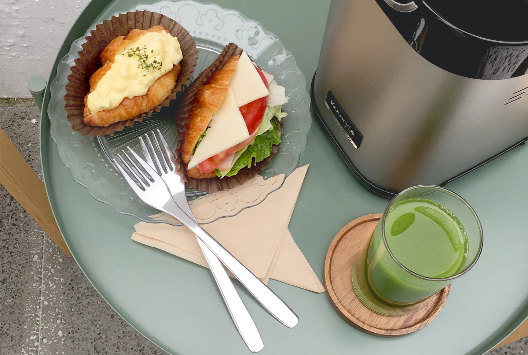 High-angle overhead view of a breakfast setting on a sage green round table, featuring a clear glass of fresh green juice on a wooden coaster next to the silver stainless steel base of a Kuvings Chef CS700 commercial juicer, paired with a glass plate holding two filled croissants and two metal forks.