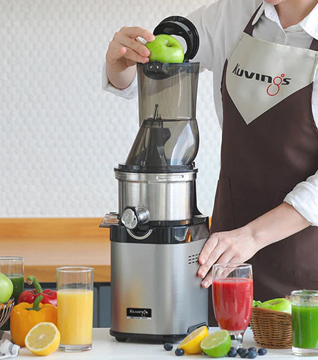 Eye-level medium shot of a person wearing a brown Kuvings apron inserting a whole green apple into the top flip-gate lid of a stainless steel Kuvings Chef CS700 commercial slow juicer, surrounded by glasses of assorted fruit juices and fresh produce on a countertop.