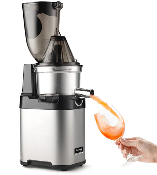 Three-quarter product shot of a stainless steel Kuvings Chef CS700 commercial slow juicer with its wide-mouth flip-gate feeding chute lid attached, showing a hand tilting a wine glass to catch fresh pink grapefruit juice pouring out of the open metal spout against a white background.