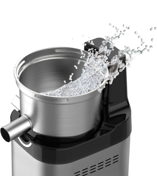 High-angle close-up studio product shot of the brushed stainless steel juicing bowl component mounted on the base of a Kuvings Chef CS700 commercial slow juicer, dynamically showing a clean water splash erupting from the open top to illustrate ease of cleaning, set against a solid white background.