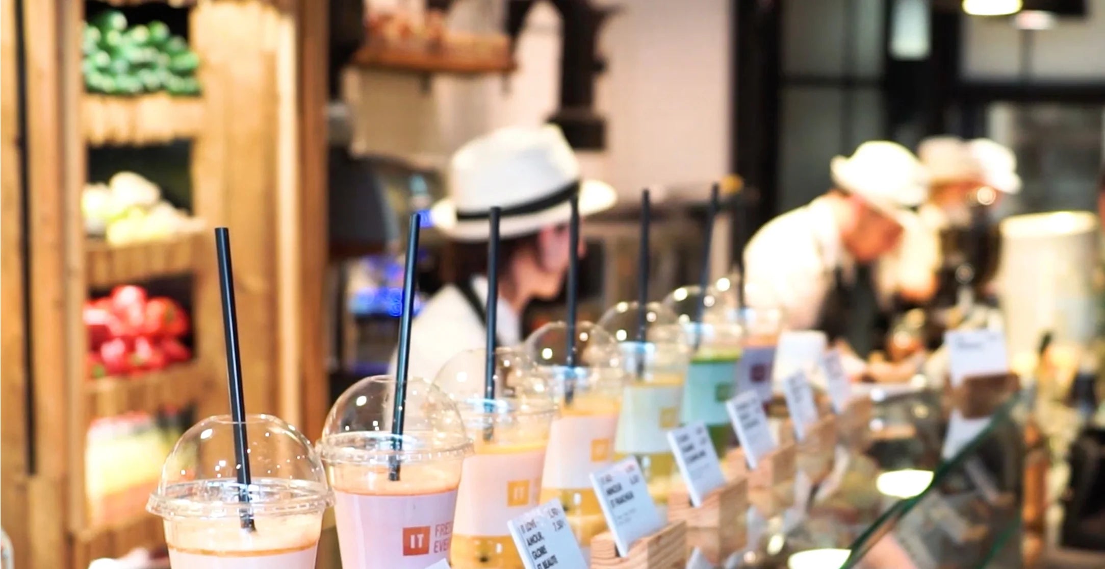 Eye-level, shallow depth-of-field shot of a busy juice bar counter showing a row of fresh juices and smoothies in clear plastic cups with straws lined up in the foreground, while staff members in matching white hats work out of focus in the background.