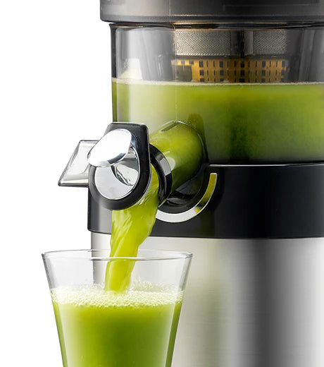Straight-on, close-up studio shot of the chrome smart cap spout of a Kuvings CS600 Chef slow juicer open, pouring fresh, vibrant green juice into a clear glass against a white background.