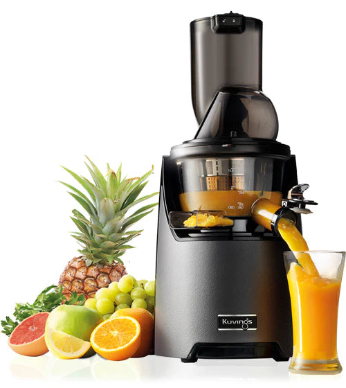 Three-quarter studio product shot of a gunmetal grey Kuvings EVO820 whole slow juicer actively dispensing bright orange juice into a tall clear glass, beautifully arranged alongside fresh ingredients including a whole pineapple, green grapes, a green apple, orange halves, and a grapefruit slice against a crisp white background.