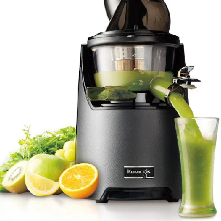 Three-quarter studio product shot of a gunmetal grey Kuvings EVO820 whole slow juicer actively dispensing vibrant green juice into a clear glass, arranged with fresh ingredients including an orange half, lemon and kiwi slices, a green apple, and leafy green vegetables against a crisp white background.
