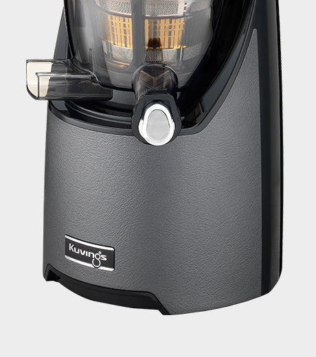 Three-quarter close-up studio product shot of the lower section of a Kuvings EVO820 whole slow juicer, highlighting its textured gunmetal graphite grey motor base with a black Kuvings logo badge, assembled with a dark translucent juicing bowl and closed chrome smart cap against a light grey background.