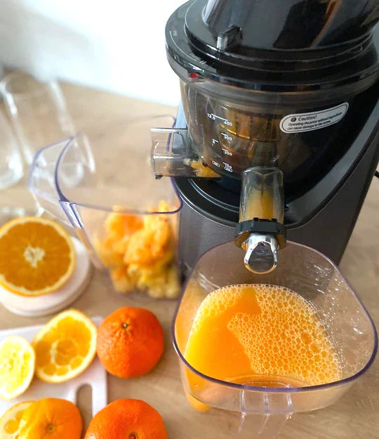 High-angle close-up lifestyle shot of a dark grey Kuvings EVO820 whole slow juicer actively pressing citrus fruit, showing fresh orange juice with foam filling the clear plastic juice pitcher on a wooden table, surrounded by tangerine halves, whole oranges, and extracted pulp accumulating in the background container.