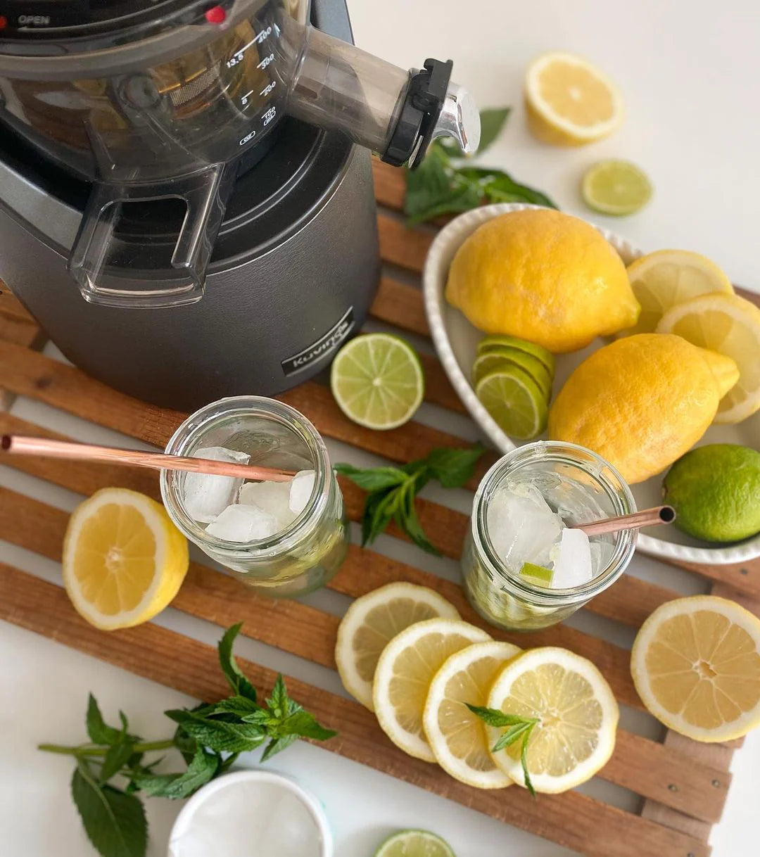 High-angle overhead lifestyle shot of a gunmetal grey Kuvings EVO820 whole slow juicer positioned on a wooden slatted mat next to two glass jars filled with ice, lemonade, and copper straws, beautifully surrounded by fresh whole lemons, limes, citrus slices, and scattered mint sprigs.