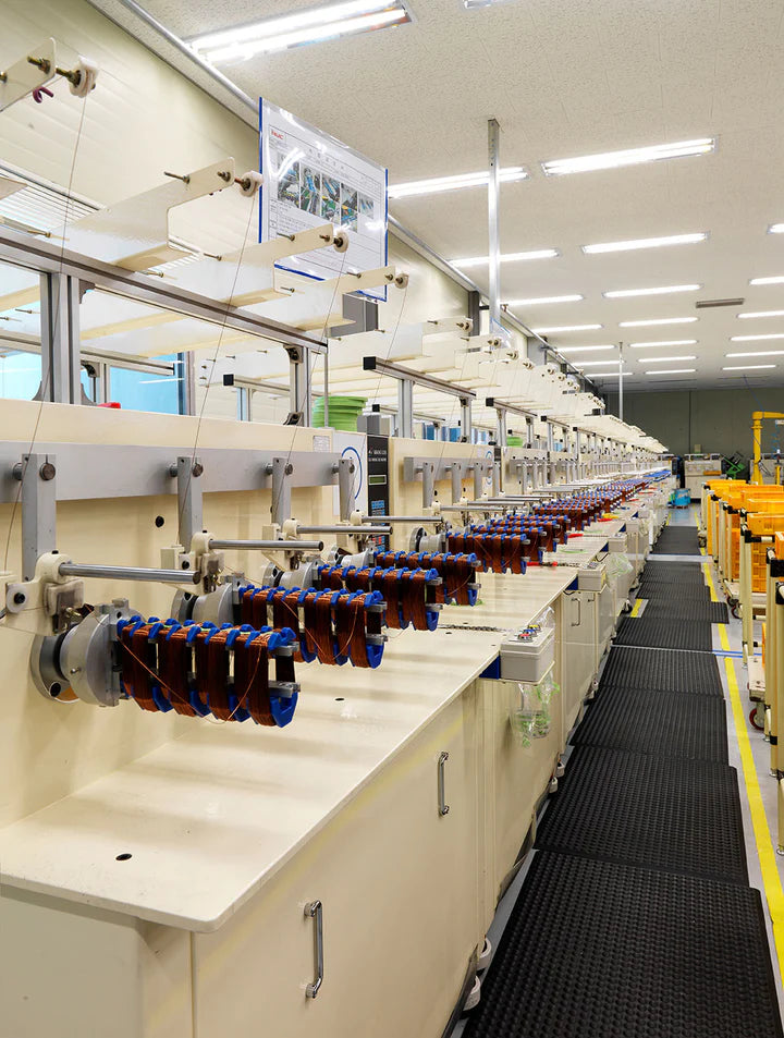Eye-level, vanishing-point perspective shot of an automated assembly line inside a well-lit Kuvings factory, showing specialized machinery winding copper wire coils for electric appliance motors stretching down a long production floor.