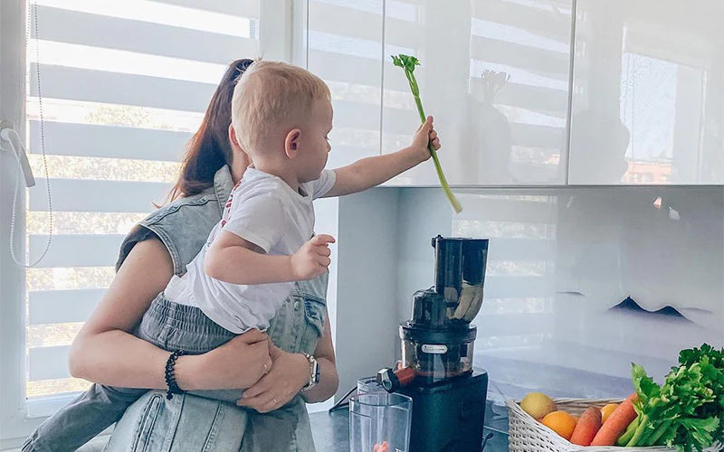 Woman holding a child in a kitchen with a kuvings coldpress juicer and fruits on the counter.