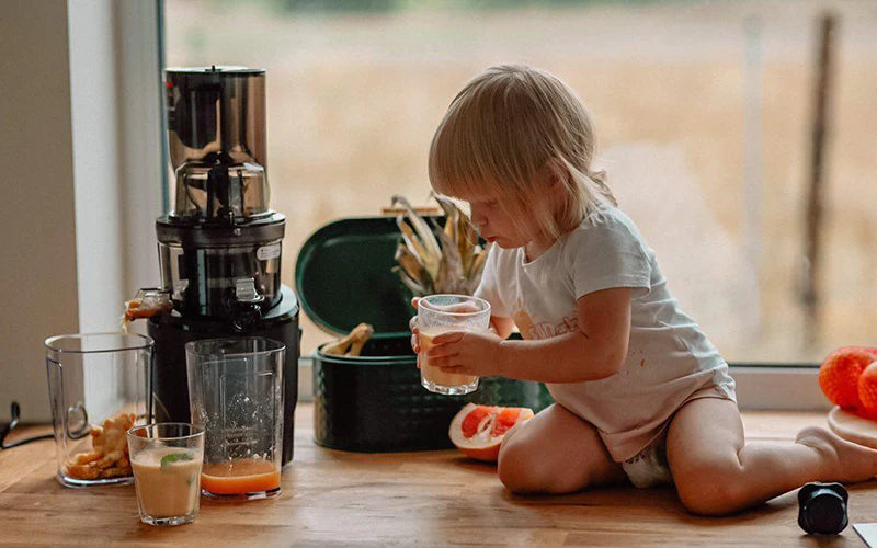 Child drinking from a glass next to a kuvings revo juicer and fruits on a wooden floor.