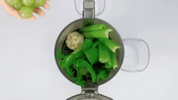 Juicer with green vegetables and a hand holding grapes on a white background