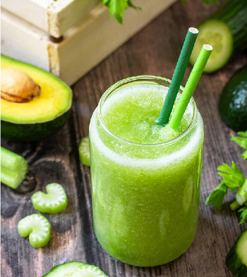 Green smoothie in a glass with a straw, surrounded by fresh vegetables on a wooden surface