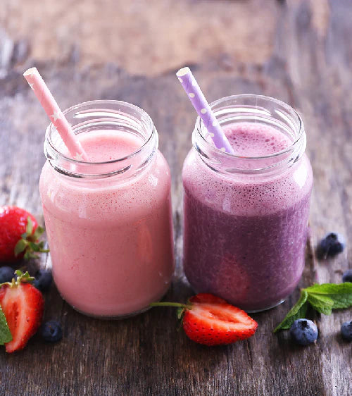 Two smoothies in glass jars with straws on a wooden surface, surrounded by berries.