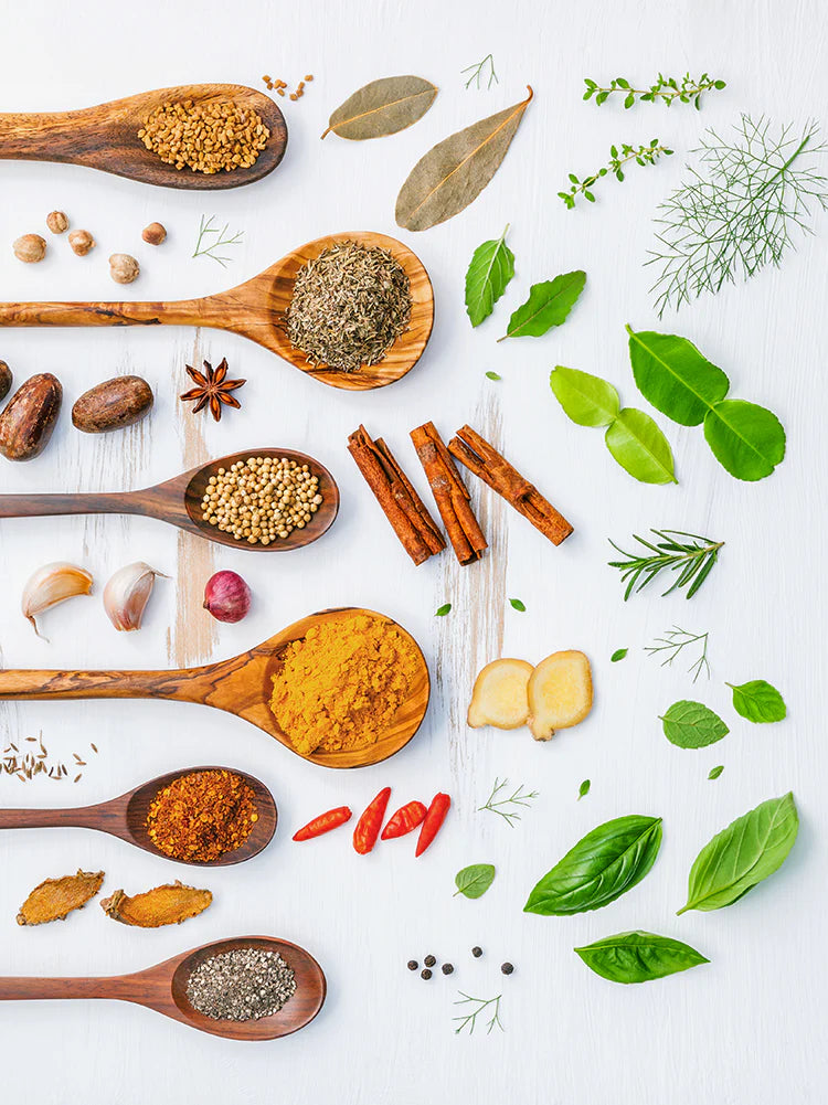 High-angle overhead studio shot of various culinary ingredients arranged on a white wooden background, featuring an assortment of dry spices, seeds, and powders in wooden spoons stacked vertically down the left side, interspersed with fresh green leaves, herbs, ginger slices, and cinnamon sticks scattered on the right.