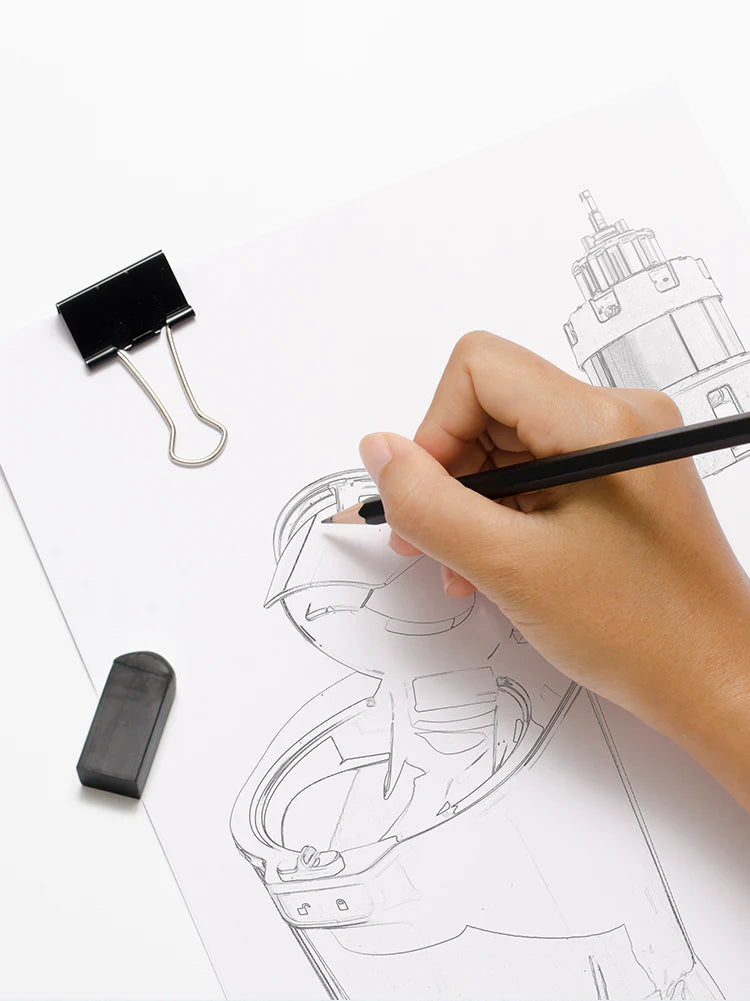 High-angle close-up studio shot of a designer's hand using a black pencil to draw a detailed product schematic sketch of the Kuvings REVO830 slow juicer feeding chute and flip-gate mechanism on white paper, alongside a black binder clip and eraser.